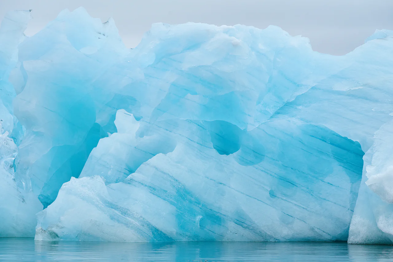 A wall of glacial ice, pale and deep blue in bands, standing in still grey water